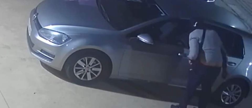 Security camera footage showing a person breaking into a car in a car park at night.