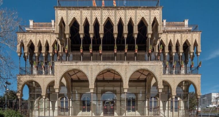 A historic Neo-Mudéjar building, Casa de les Altures, with intricate brickwork and archways, surrounded by green trees under 