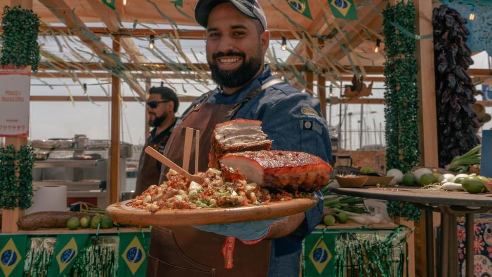 A chef grilling meat over an open fire at a food festival, with smoke rising and people in the background.