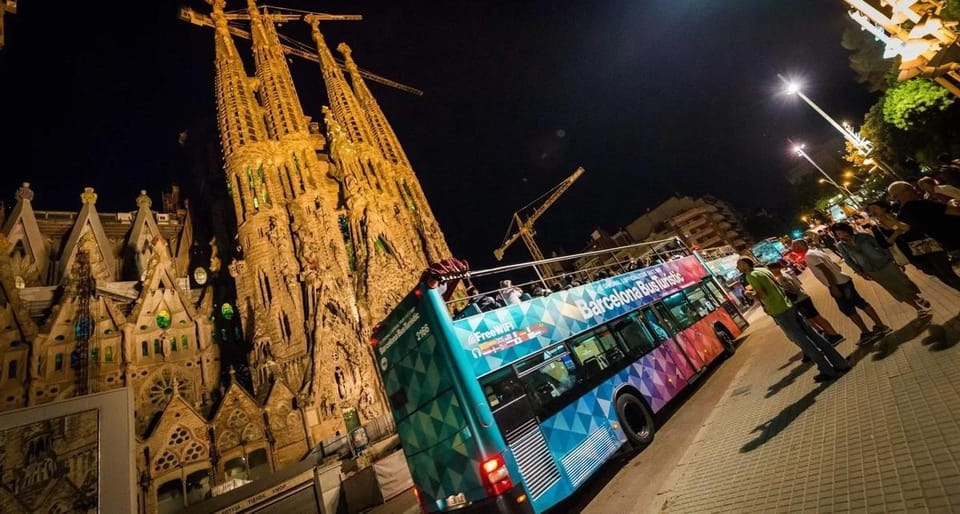 An open-top double-decker Barcelona Night Tour bus driving past illuminated landmarks at night, with passengers on the upper 