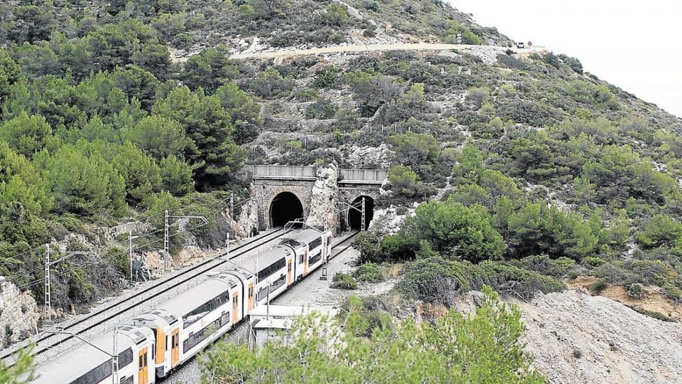 Rodalies train travelling through a tunnel near the coast, with blue sky and sea visible outside.