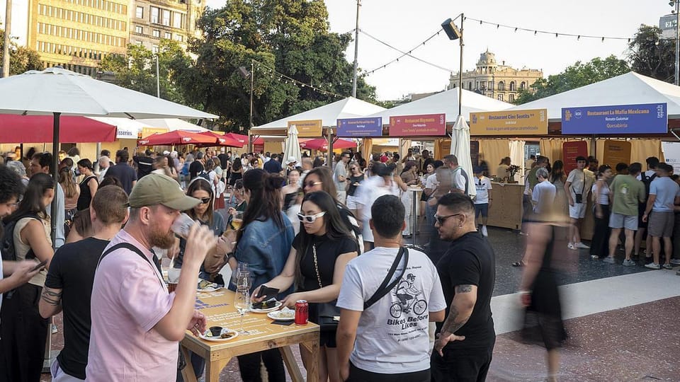 People enjoying food and drinks at outdoor stalls in Plaça Catalunya, Barcelona, during a gastronomic fair.