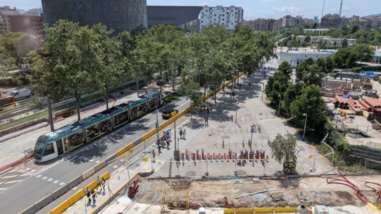 A modern tram travelling along a street in Barcelona, with buildings and trees in the background under a clear sky.