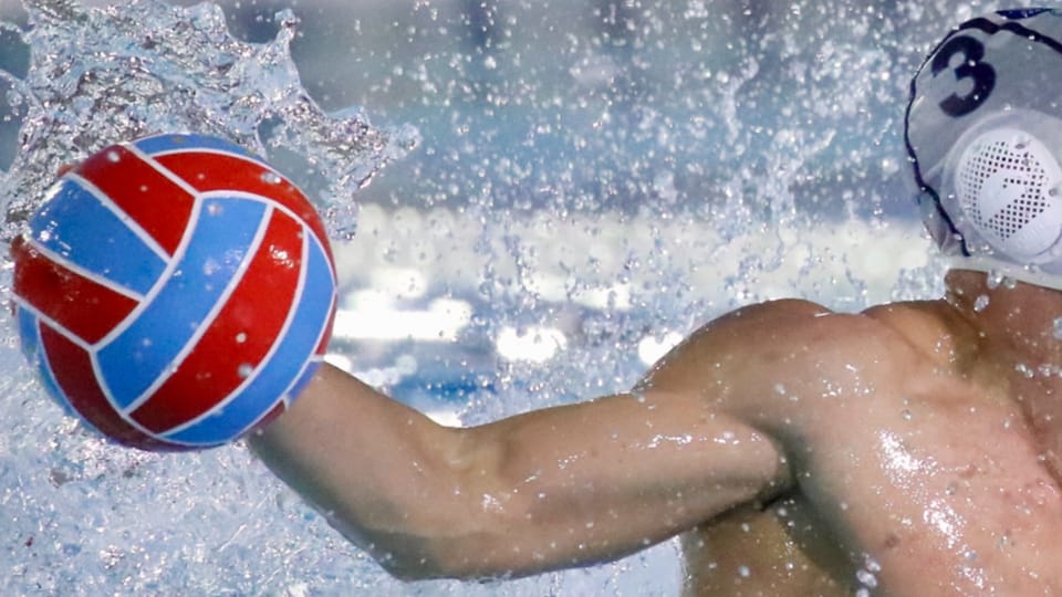 A water polo player in a dark cap throws the ball during a match, with other players in the water and spectators in the backg